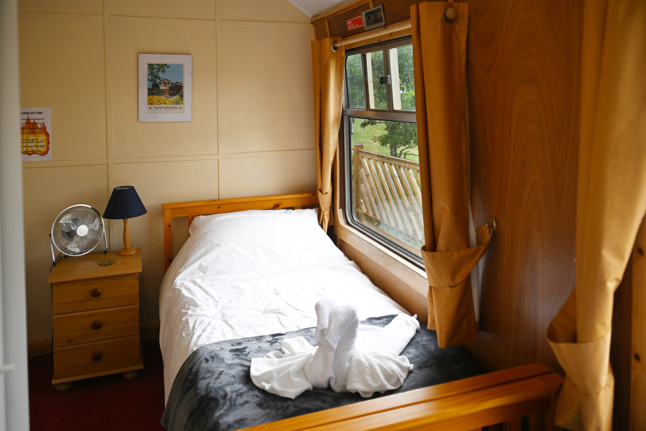 1 of two single beds found in the second room. It is under a window with curtains, a bedside table is beside it with a lamp and fan.